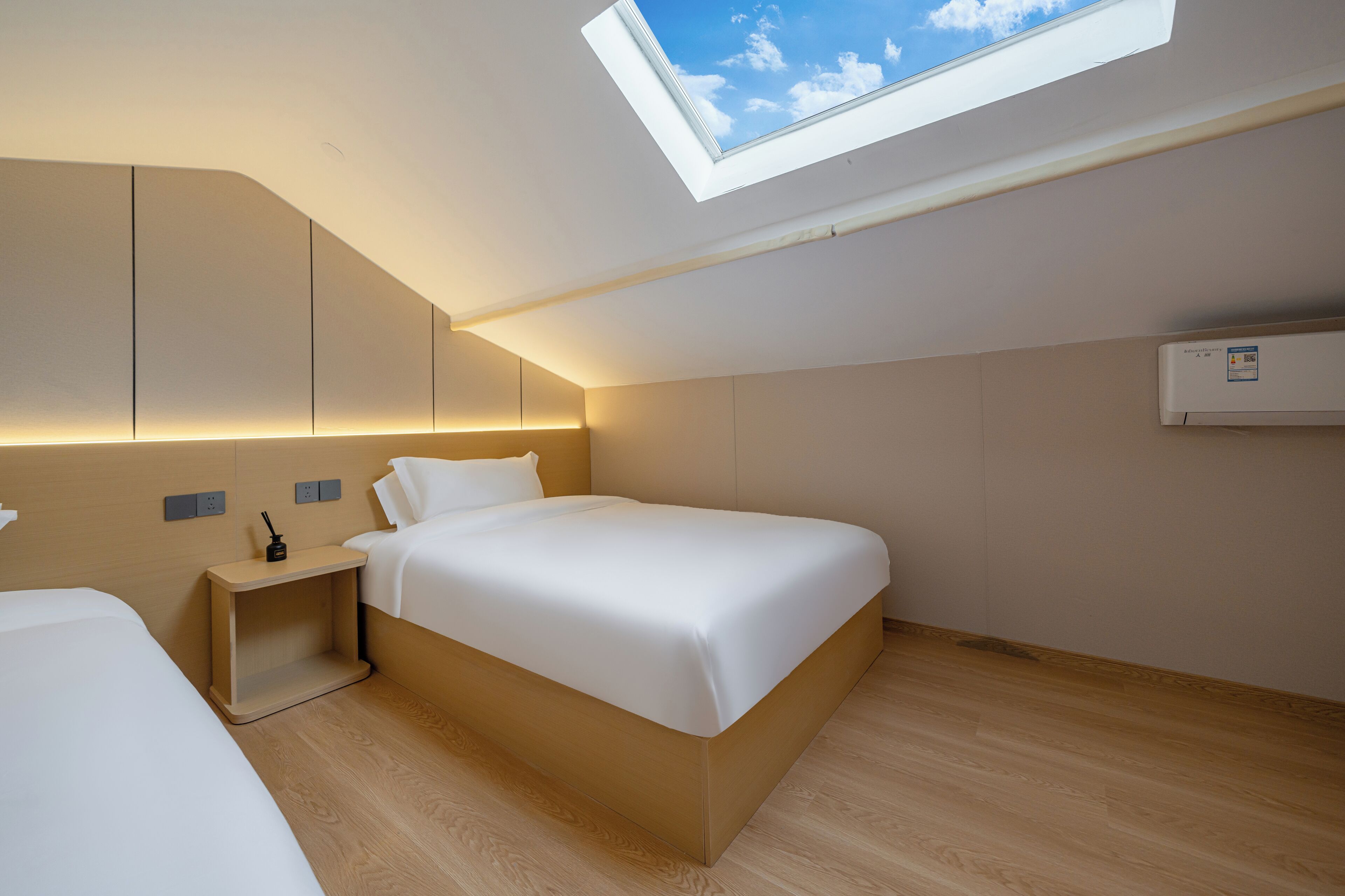 A modern bedroom with a skylight, a bed, bedside tables, and a wall-mounted air conditioning unit.