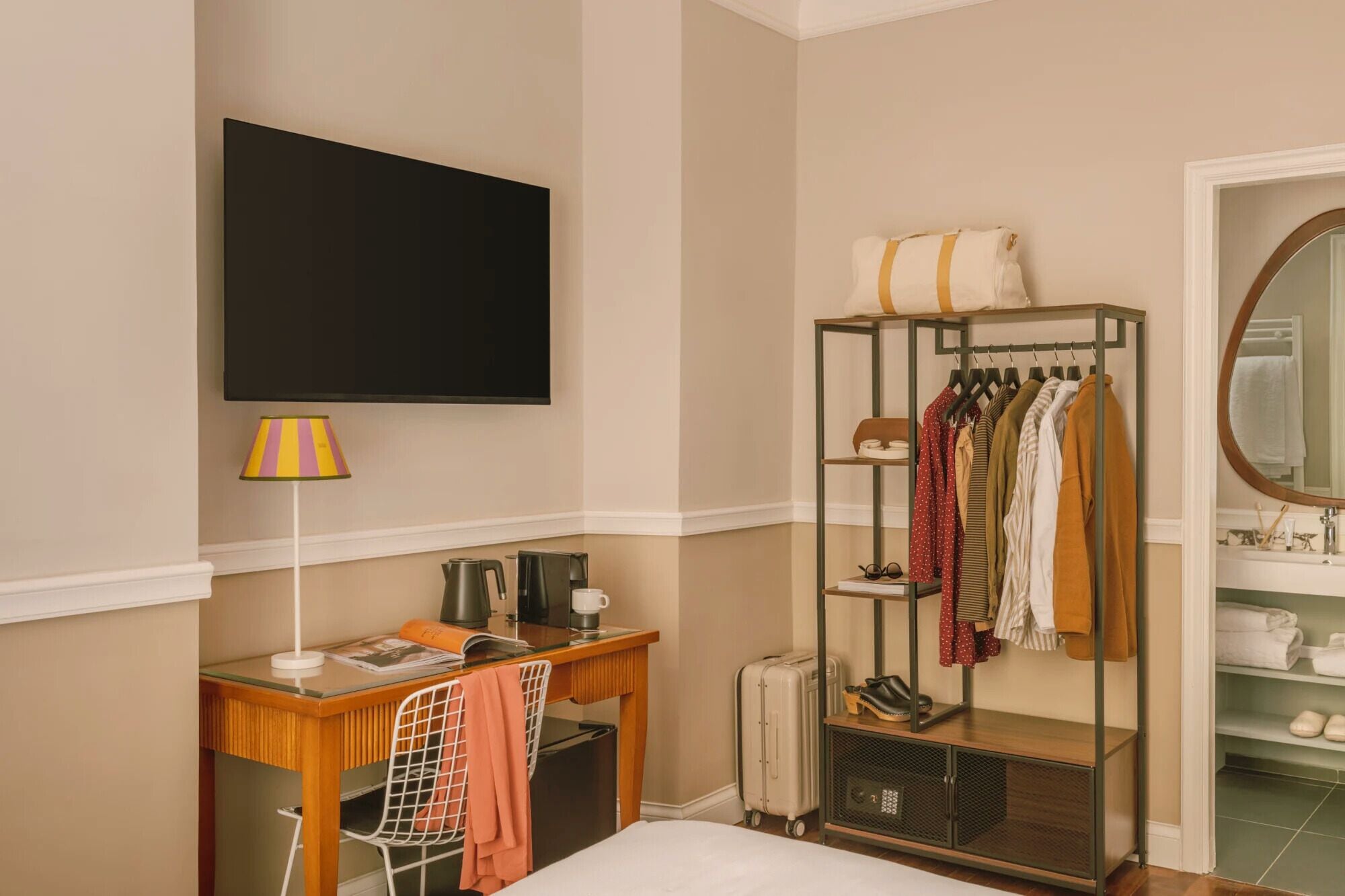 Een hotelkamer met een flatscreen televisie aan de muur, een bureau met een lamp, een kapstok met kleding en een koffer.