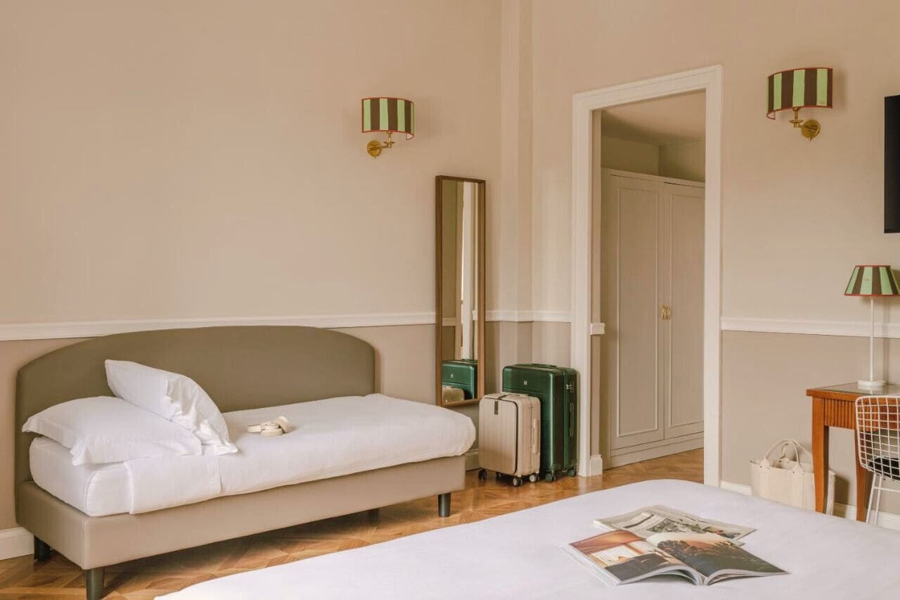 Habitación de hotel con cama, sofá, espejo, armario y escritorio con silla. Hay dos maletas y un bolso en el suelo.