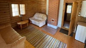 Un acogedor interior de cabaña con techo de madera, chimenea de ladrillo, sofá de cuero blanco y una pequeña mesa.