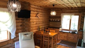 Interior acogedor de una cabaña de troncos con comedor, un sillón blanco, un televisor montado en la pared y una cocina con armarios de madera y un ventanal.