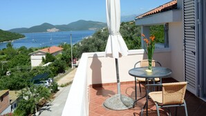 Een balkon met een tafel en stoelen, een witte parasol en uitzicht op zee en bergen.