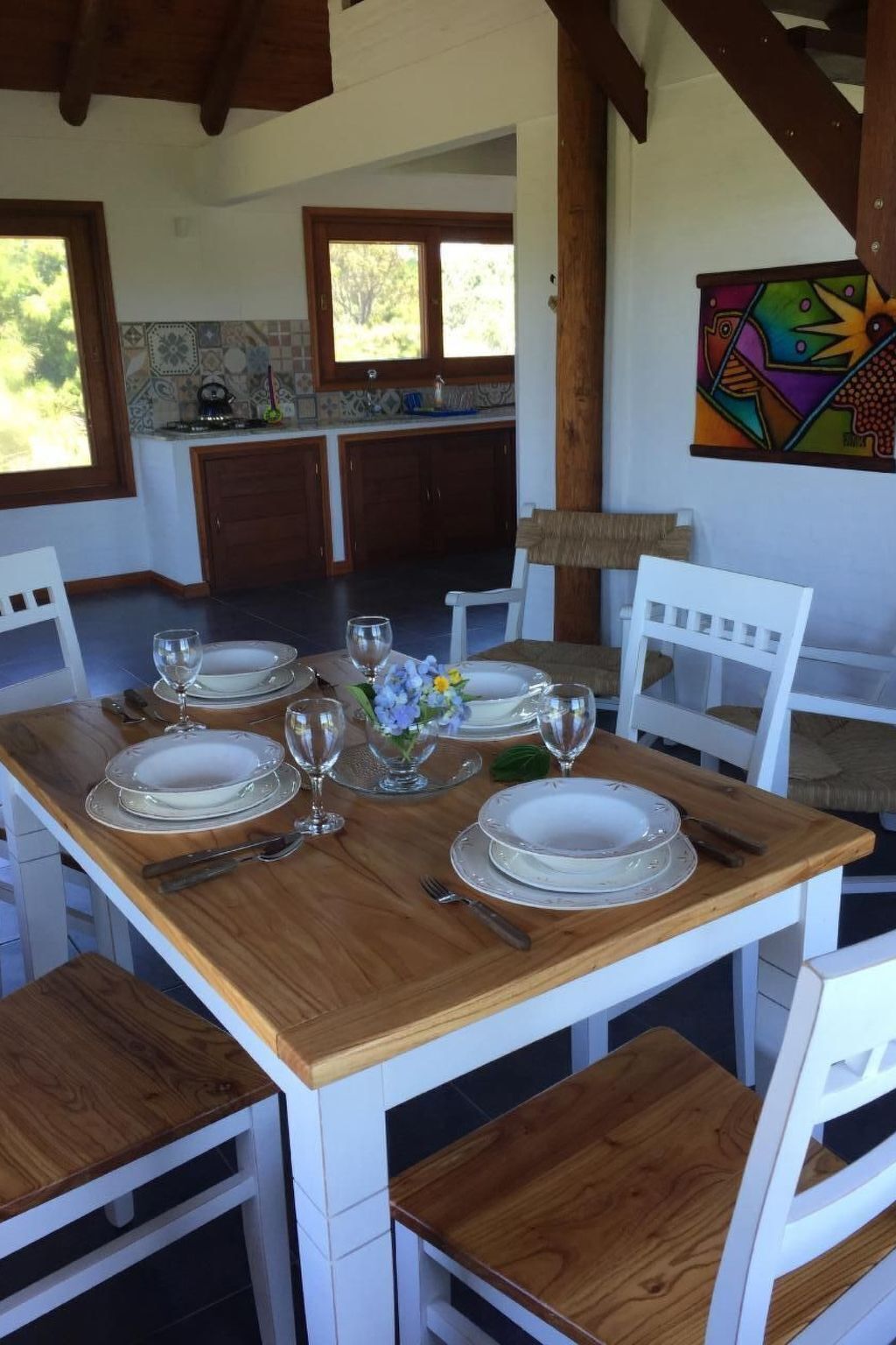 Un coin repas avec une table en bois dressée pour quatre personnes, des chaises blanches et une cuisine en arrière-plan.