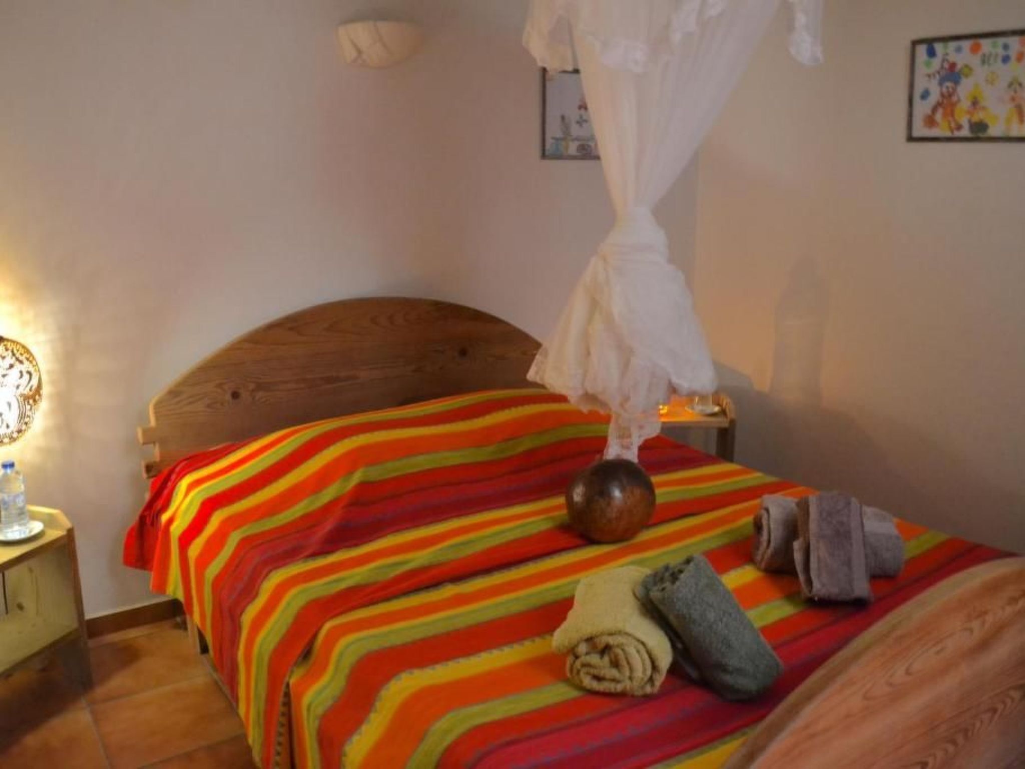 Une chambre avec une tête de lit en bois, une couvre-lit à rayures, un baldaquin blanc, une bouteille d’eau et des serviettes sur le lit.