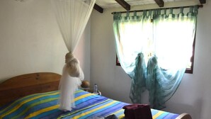 Un dormitorio con una cama, cabecera de madera, mosquitera, colcha a rayas, ventana con cortinas y una botella de agua en la mesita de noche.