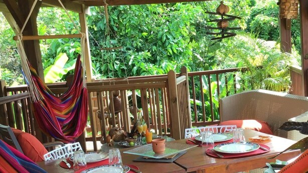 Una terraza de madera con una hamaca, mesa de comedor con platos, vasos y un cuenco de frutas.