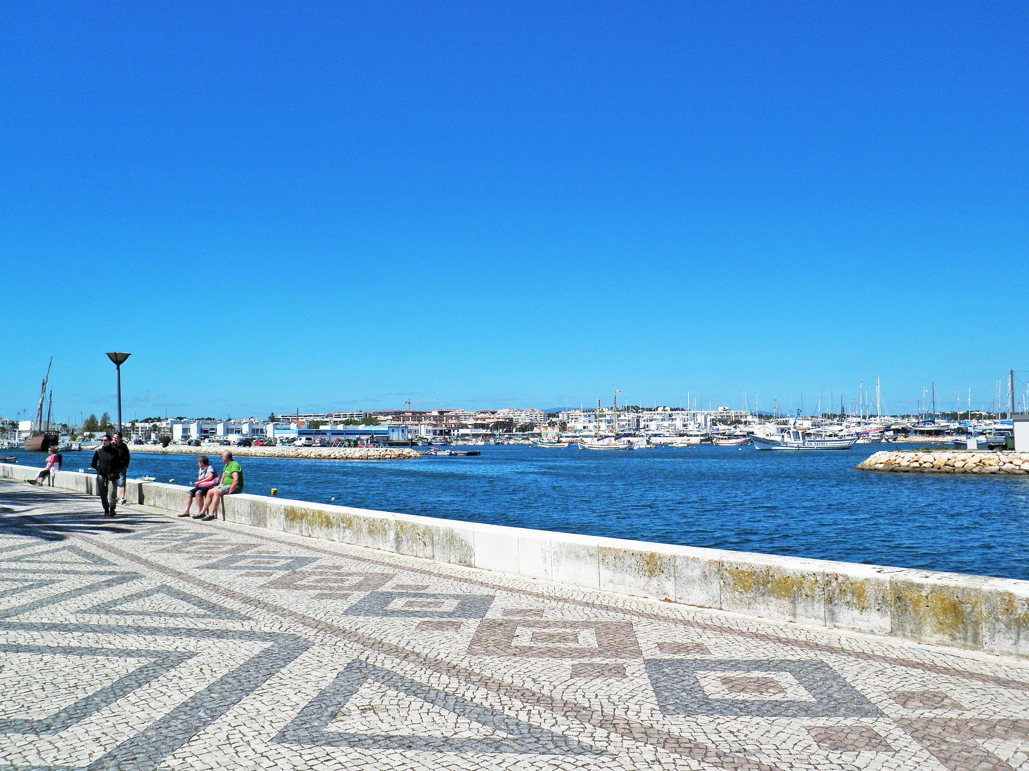Eine Uferpromenade mit Fliesenbelag und Blick auf Boote und Gebäude in der Ferne.