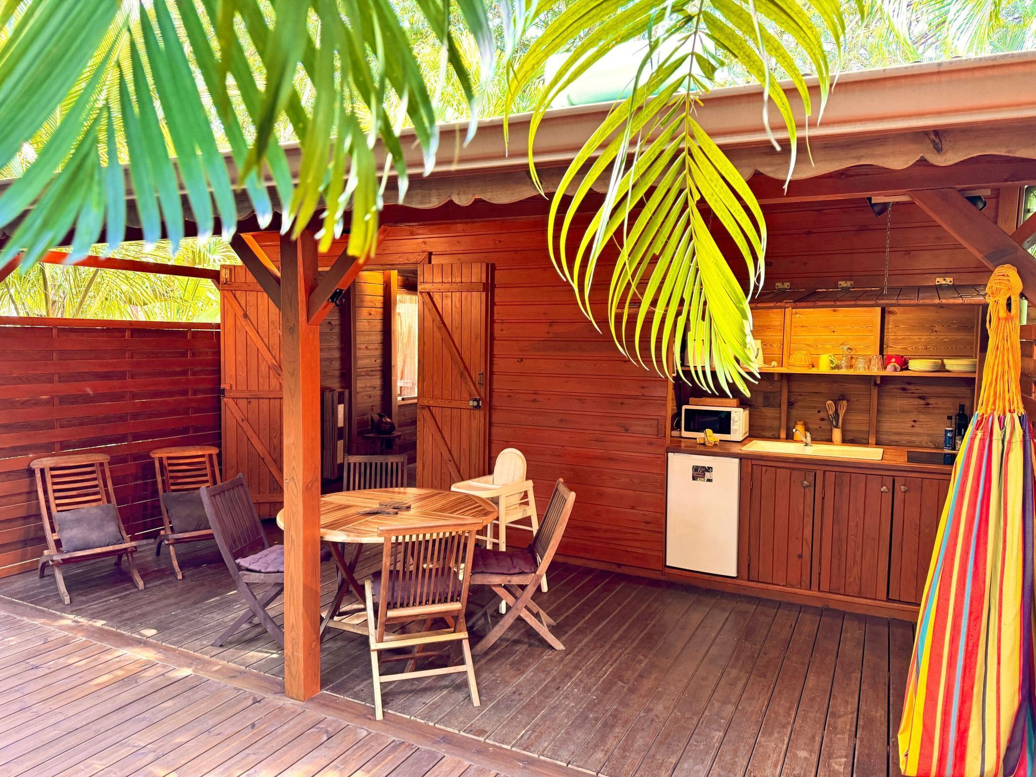 Um deck de madeira com mesa e cadeiras, uma pequena cozinha com geladeira e micro-ondas, e uma planta pendurada.