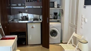 A compact kitchen with a refrigerator, microwave, and washing machine. There is a small dining area with a table and chairs. - Naousa Center Living (Paros)