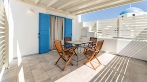 A covered outdoor area with a table and chairs, a blue door, and a white building with a blue dome in the background. - Naousa Center Living (Paros)