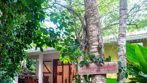 A wooden sign reading "Rouge Coral" in a lush, green outdoor area with a wooden deck and lounge chairs.