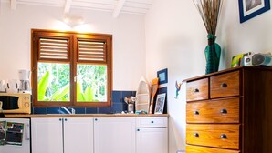 A kitchen with white cabinets, a wooden cabinet with drawers, a window with shutters, and a small trash can.
