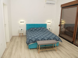 A bedroom with a teal bed, a bench, two bedside tables, and a sliding glass door.