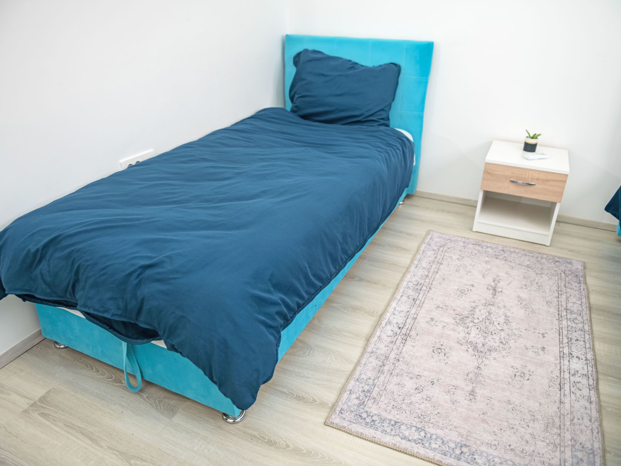 Ein Einzelbett mit petrolfarbenem Kopfteil und Fußteil, ein kleiner Nachttisch mit Schublade, ein kleiner Topfpflanze und ein gemustertes Teppich auf Holzdielen.