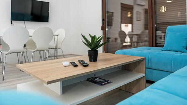 A modern living room with a wooden coffee table, a potted plant, a blue sofa, and a flat-screen TV mounted on the wall.