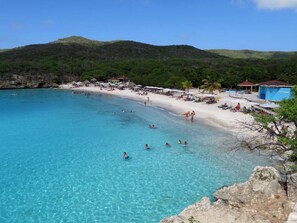 En strand med klart turkis vand, en sandet kyst og et par personer, der svømmer. Der er parasoller og strandstole langs stranden, og en bygning med et blåt tag i baggrunden.