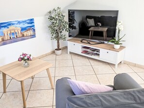 Ein Wohnzimmer mit einer Couch, einem Couchtisch, einer TV-Konsole mit Zeitschriften, einer Topfpflanze und einem Bilderrahmen mit einem Gebäude.