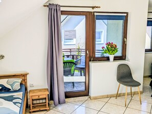Una habitación con una cama, una mesita pequeña, una silla, un ventanal con vistas al exterior y un florero con flores en el alféizar.