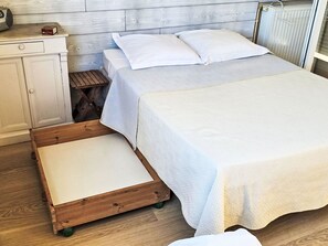 Ein Bett mit Holzbettgestell, einer weißen Matratze und einer weißen Decke. Dazu ein kleiner Holzschemel und ein weißes Schränkchen mit einem Radio.