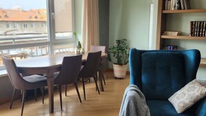 Ein modernes Wohnzimmer mit einem blauen Sofa, einem Esstisch mit Stühlen, einem Bücherregal und Blick auf Gebäude durch große Fenster.