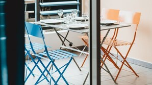 A dining area with a table set for two, blue and orange chairs, and a window with blinds. - Wohnung "Le Jardin de Corail" by Interhome (Saint-Gilles-les Bains)