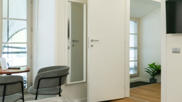 A modern hotel room with a bed, a desk, and a chair. There is a door leading to another room and a plant in the corner. - Turin Central Carlo Felice (Turin)