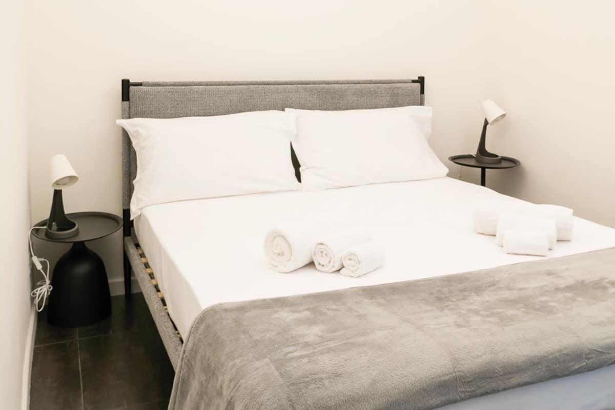 Ein ordentlich bezogenes Bett mit weißen Leinen und einer grauen Decke, zwei Nachttische mit Lampen sowie ein kleiner Tisch ebenfalls mit einer Lampe.