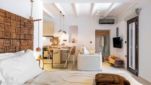 Un dormitorio con una cama grande, cabecera de madera, zona de comedor con mesa y sillas, kitchenette y puerta corrediza de vidrio que da a un balcón.