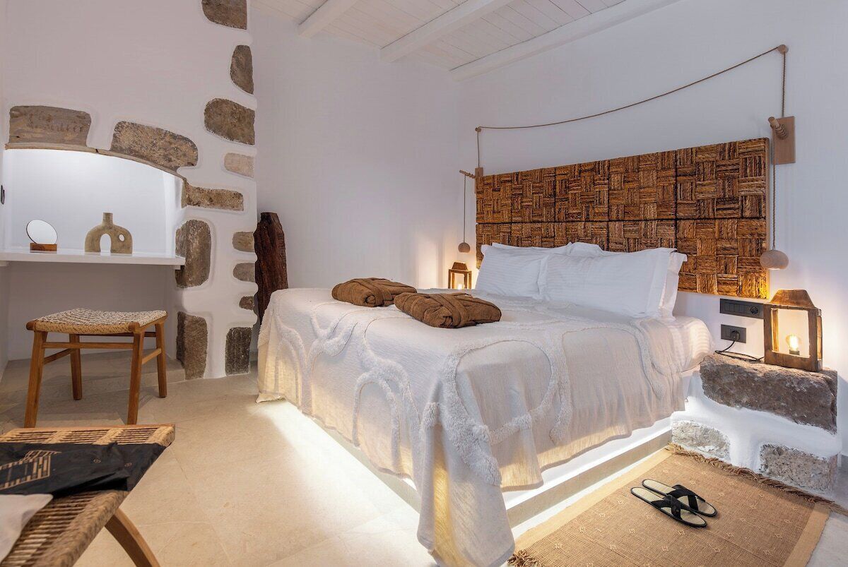 A bedroom with a stone wall, a bed with a woven headboard, a wooden chair, and a nightstand with a lamp.
