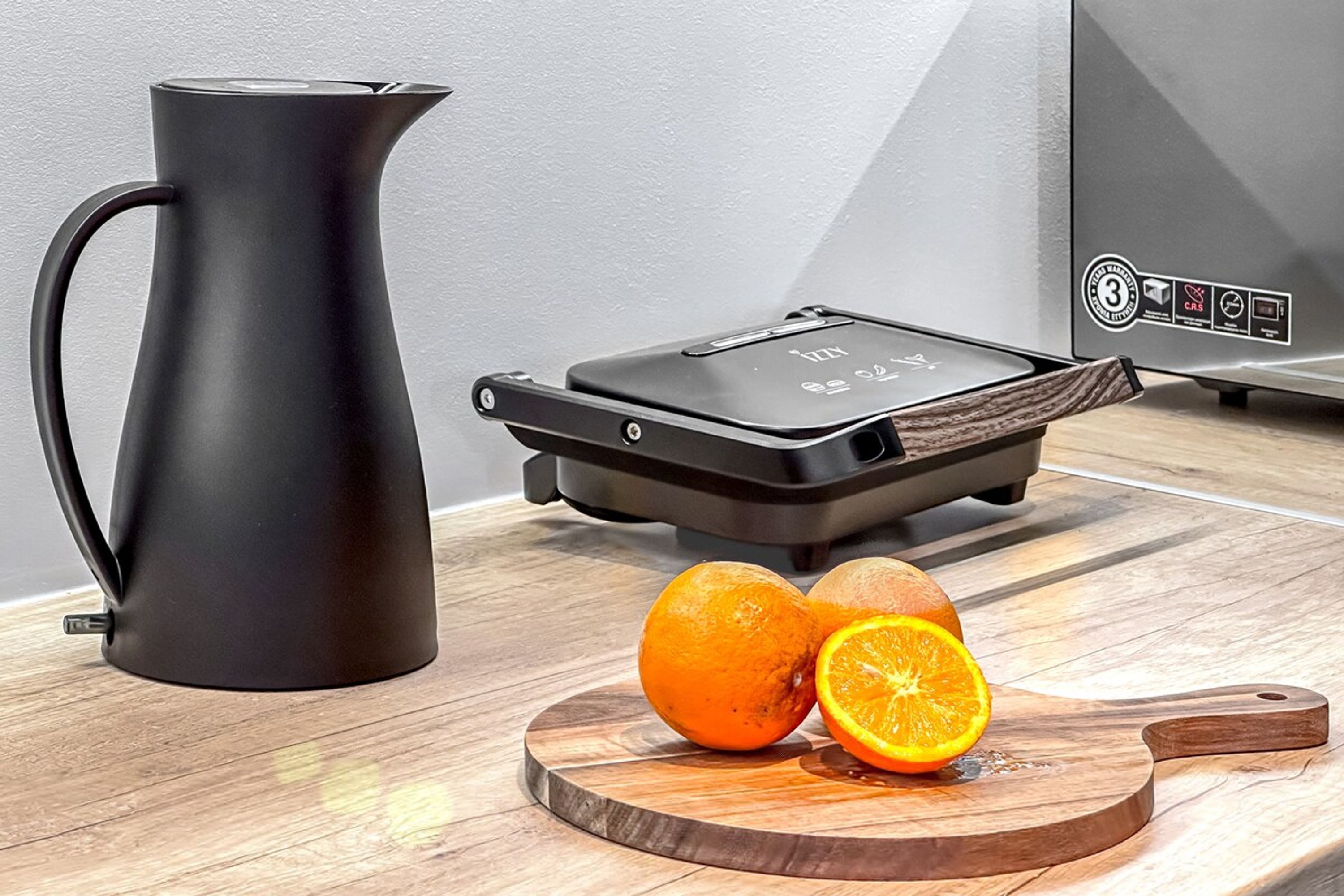 A kitchen with a black kettle, a toaster, and a wooden cutting board with oranges.