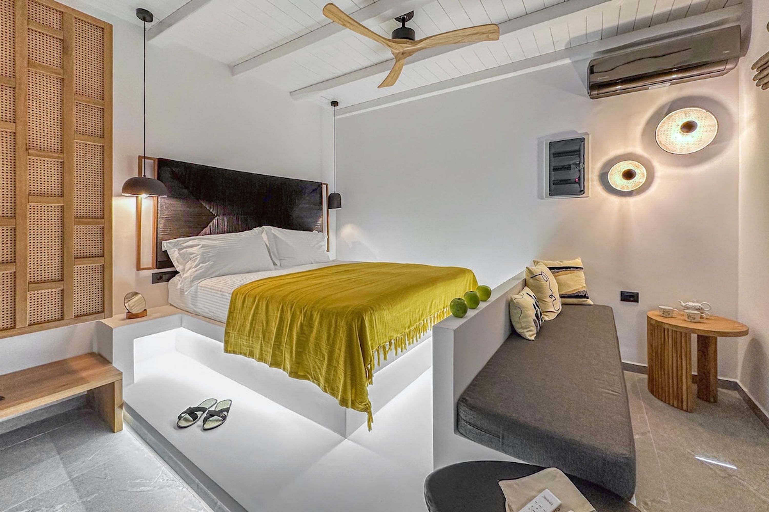 A modern bedroom with a bed, bedside table, bench, and a small wooden table with a lamp.