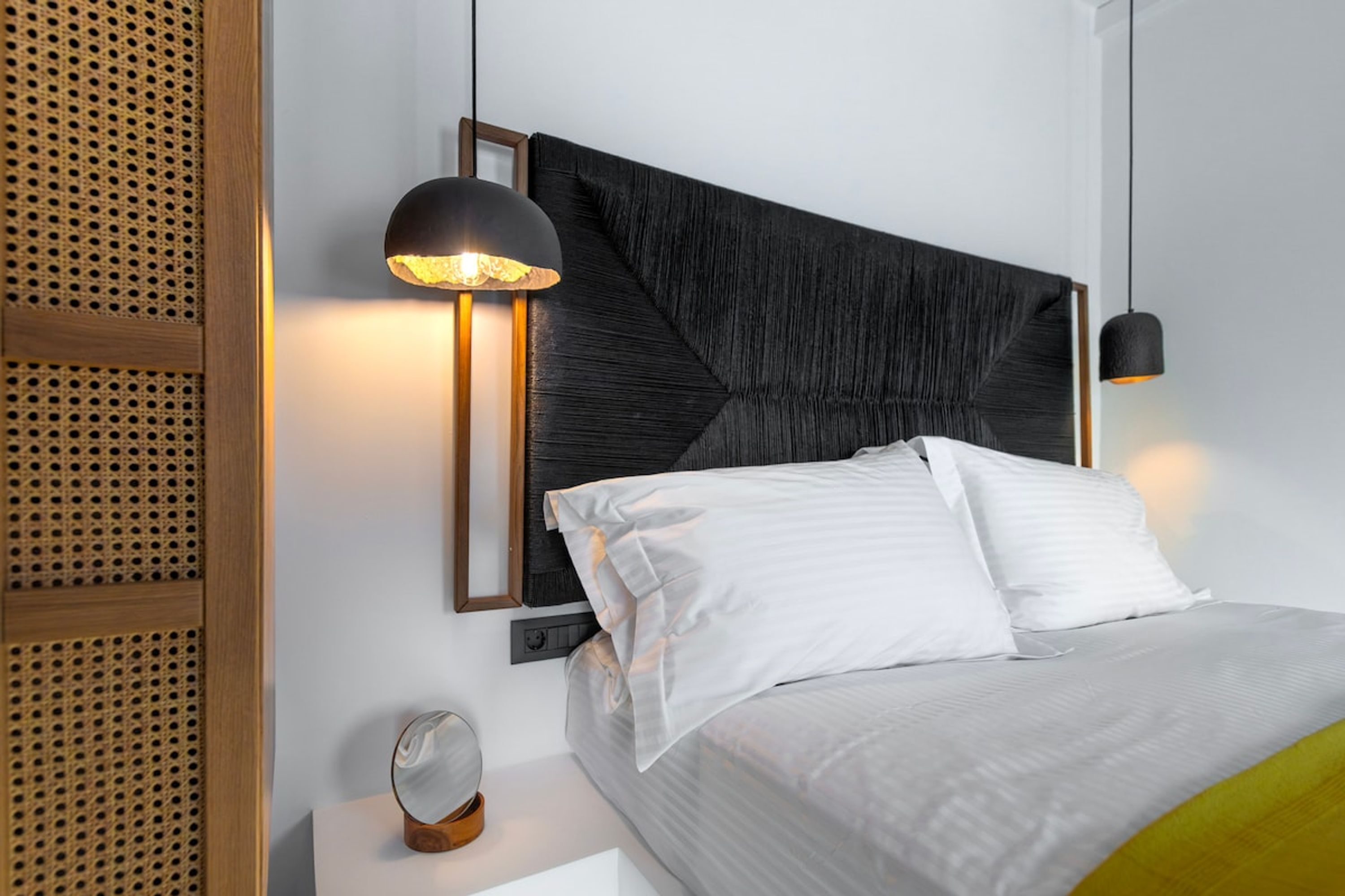 Et moderne hotelværelse med en stor seng, en sengegavl med geometriske mønstre, et natbord med et spejl og en vægmonteret lampe.