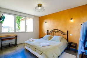 Une chambre à coucher comprenant un lit, un bureau, une chaise, une table de chevet, une lampe fixée au mur et une fenêtre donnant sur un paysage verdoyant.