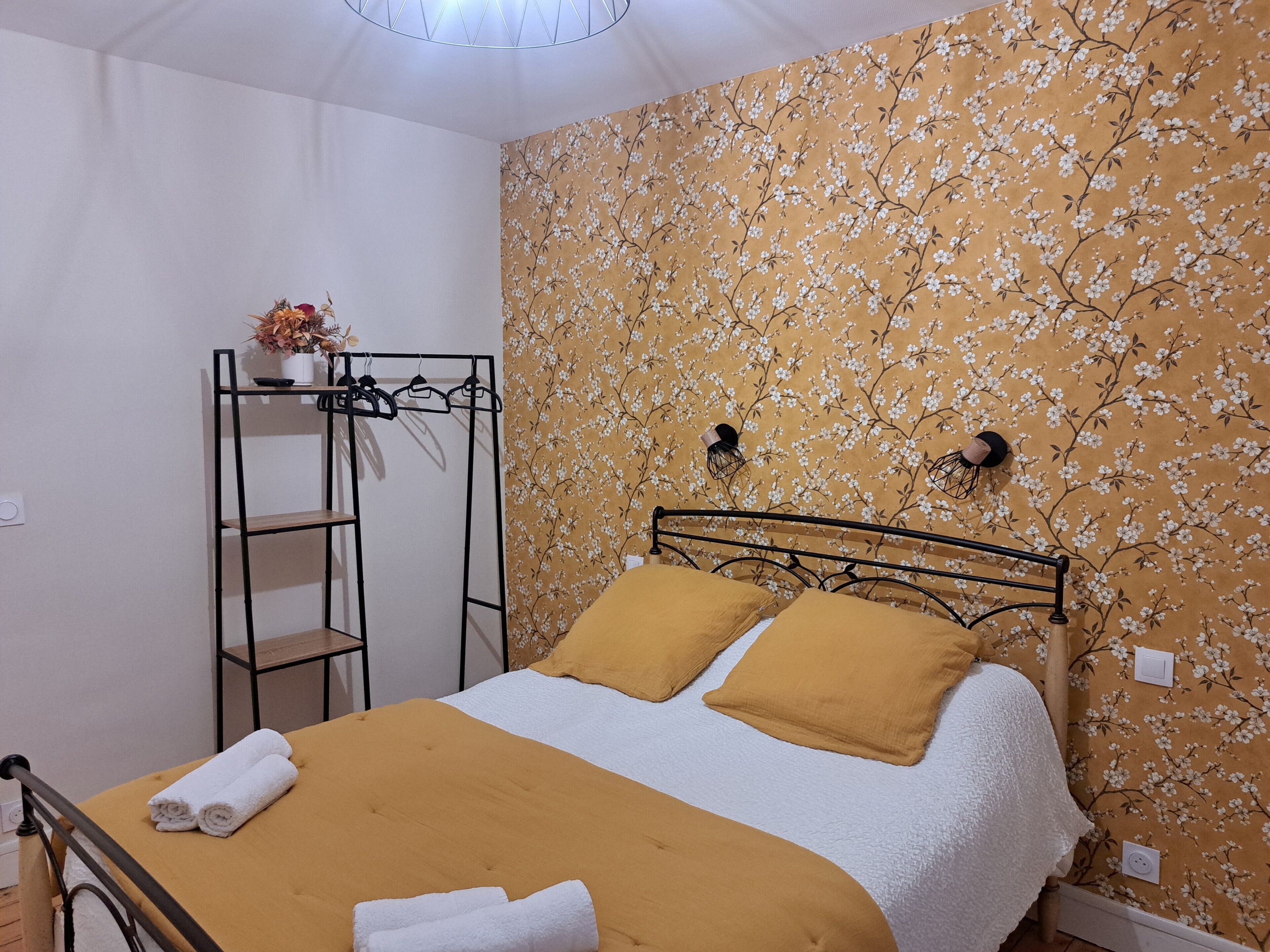 Uma cama bem arrumada com manta e travesseiros amarelos, uma estante de metal com um vaso de flores e um papel de parede com padrão floral.