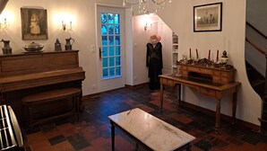 A room with a piano, a table, a bench, a chandelier, and a door with a stained glass window. - Gîte LEBASLADIN for 7 Guests – Renovated Holiday Home in Lison (Lison)