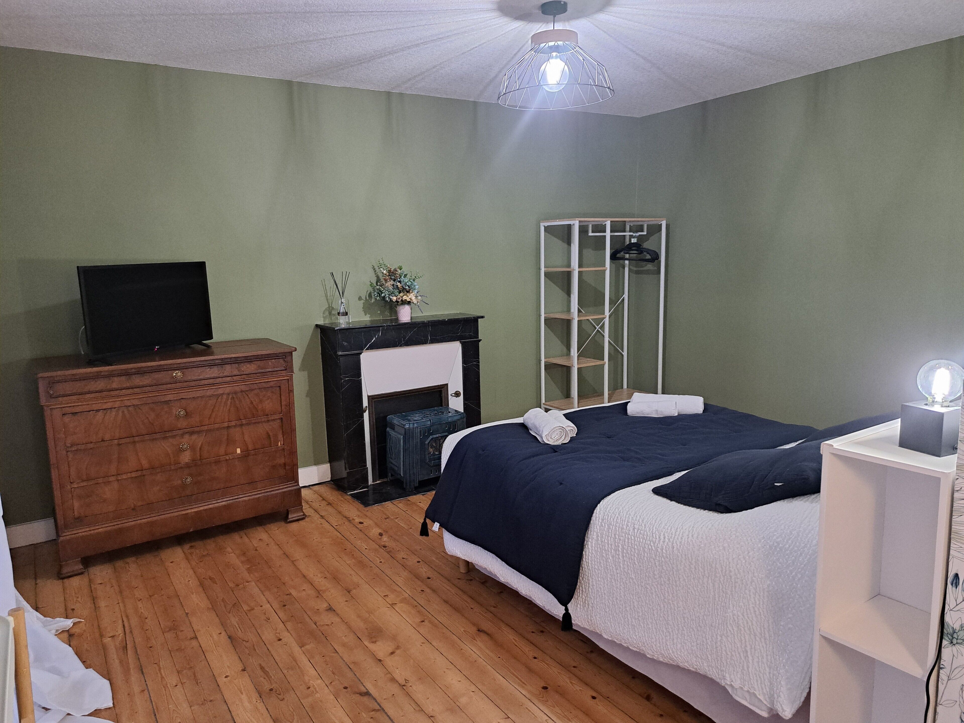 Une chambre à coucher avec une commode en bois, un téléviseur, une cheminée, un lit recouvert d’une couvre-lit blanche et bleu foncé, et un miroir avec des étagères.