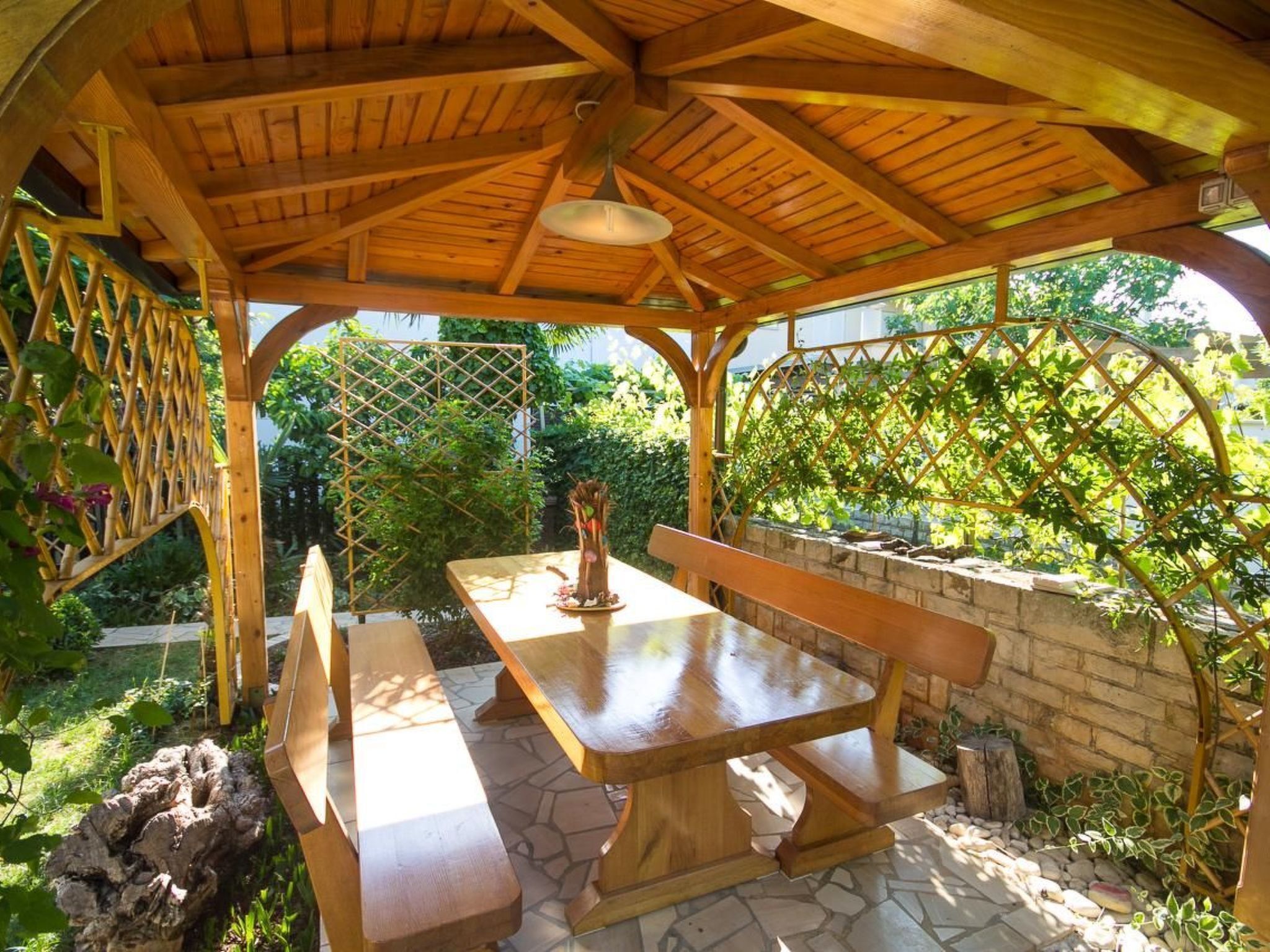 A wooden pergola with a table and benches, surrounded by greenery and stone pathways.