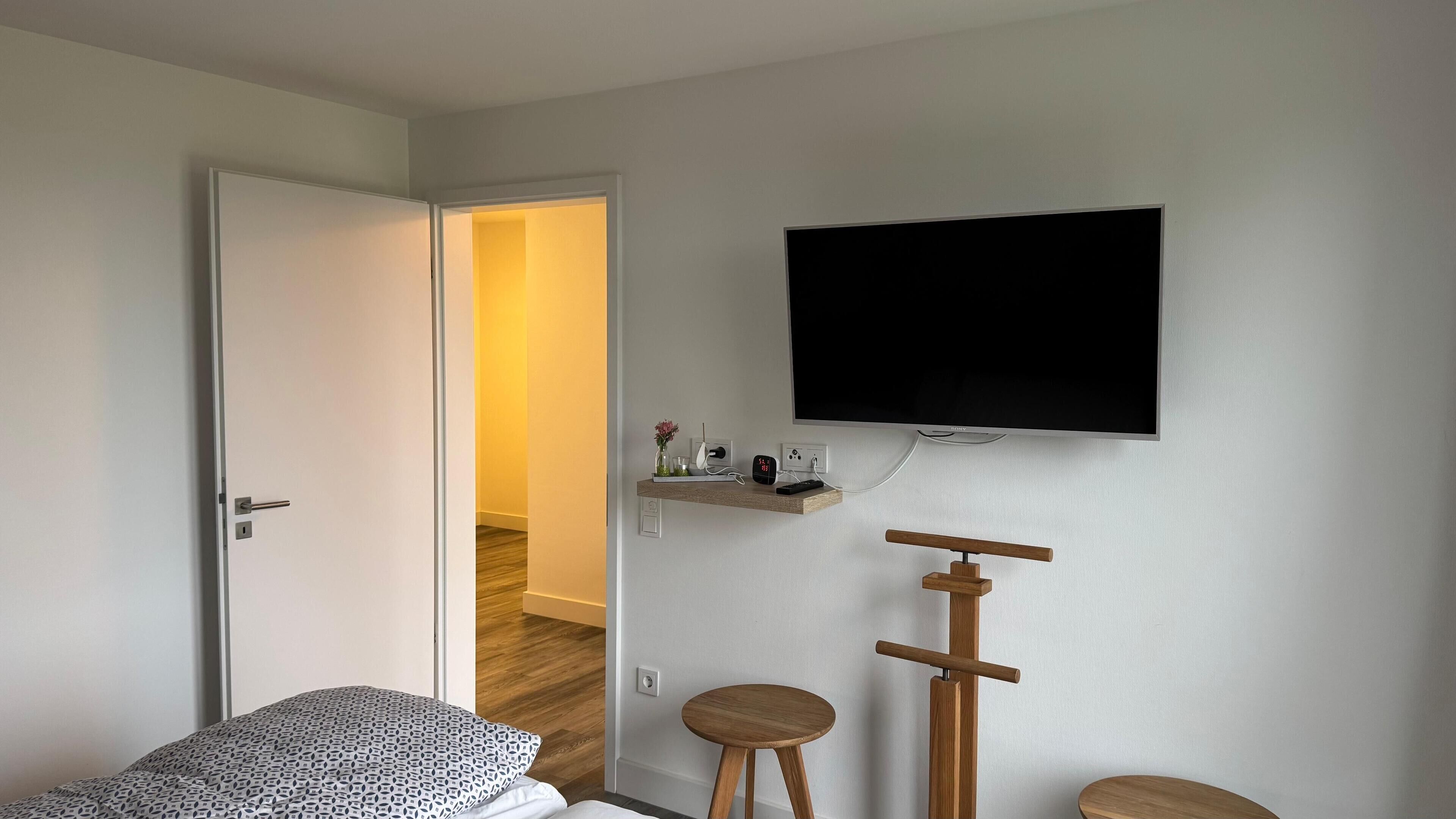 A bedroom with a bed, a TV mounted on the wall, a small wooden stool, and a bedside table with a clock.