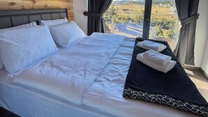 Un dormitorio con techo de madera, un ventanal con cortinas, una cama con ropa blanca y vistas a un paisaje montañoso.