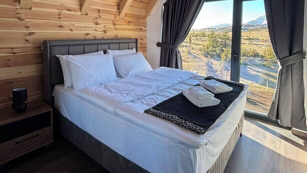 Un dormitorio con techo de madera, una cama grande, una mesita de noche y una ventana con vistas a un paisaje montañoso.