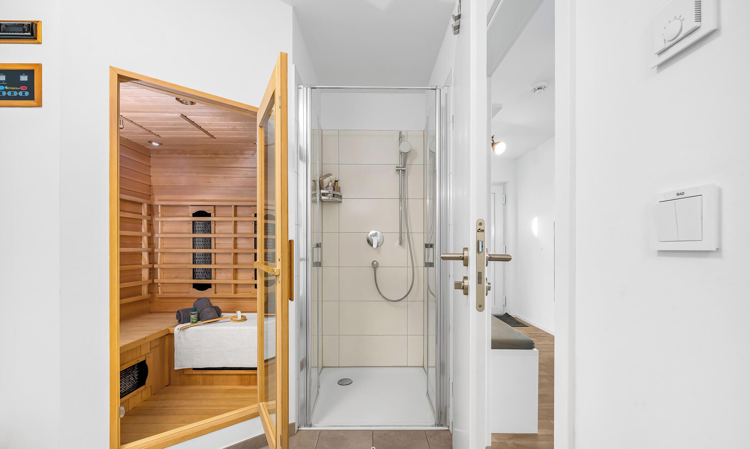 A modern bathroom with a glass shower enclosure, a wooden sauna, and a white tiled floor.
