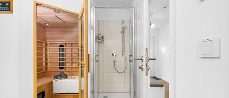 A modern bathroom with a glass shower enclosure, a wooden sauna, and a white tiled floor.