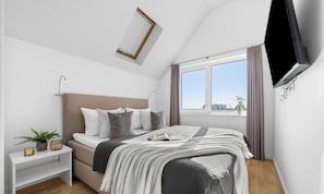Ein Schlafzimmer mit Bett, Nachttisch, Lampe, Fenster und einem an der Wand befestigten Fernseher.