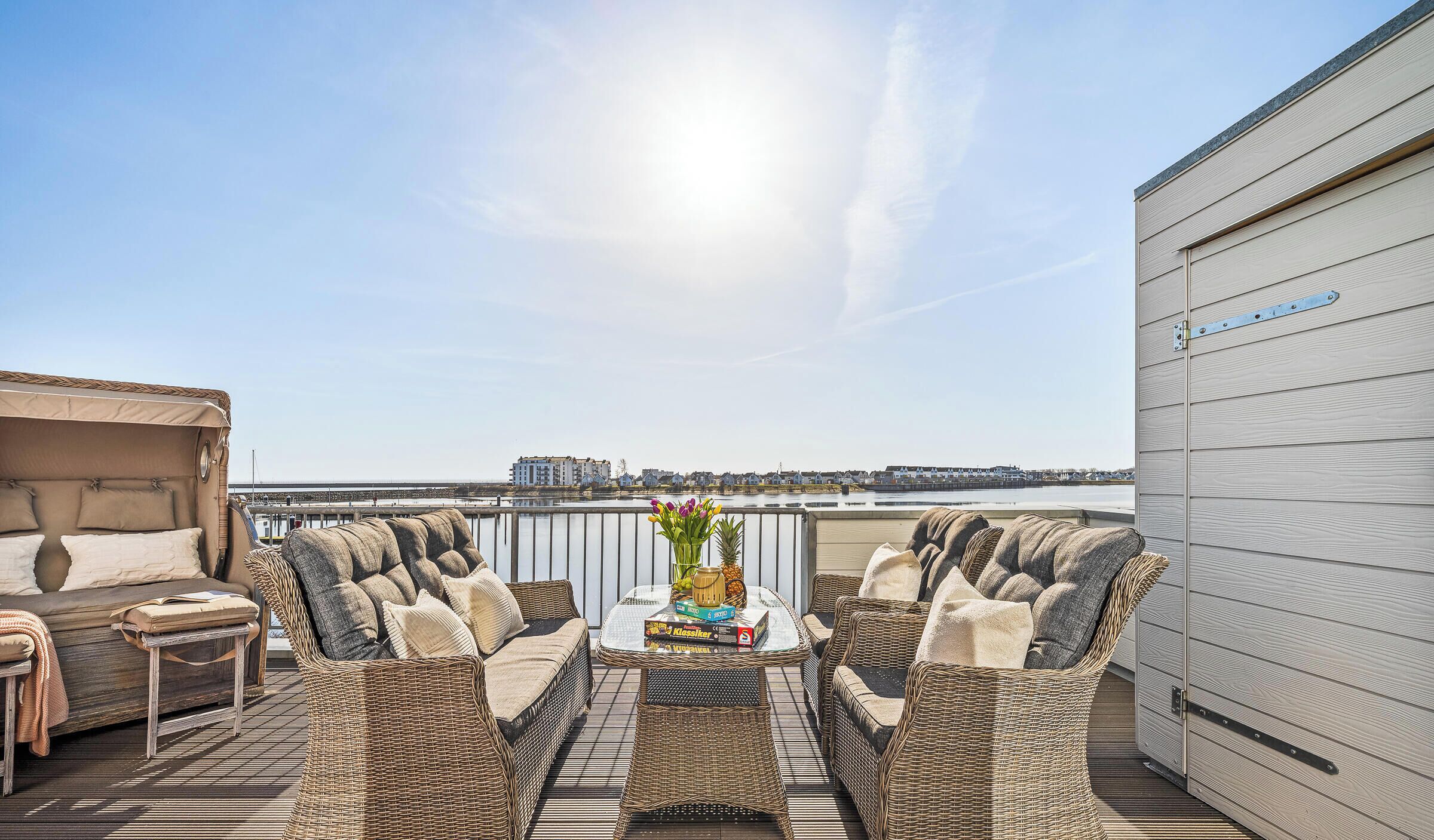 A rooftop terrace with wicker furniture, a glass table with a vase and pineapple, and a view of a waterfront.