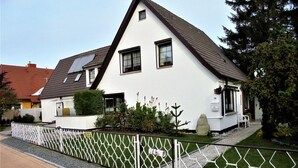 Une maison blanche à deux étages, avec un toit en pente, une cour clôturée et une allée pavée.