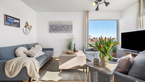 Una sala de estar moderna con un sofá gris, una mesa de centro y un televisor. Hay elementos decorativos como un jarrón con flores y un cuadro en la pared.