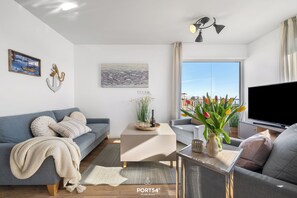 A modern living room with a grey sofa, a coffee table, and a TV. There are decorative items like a vase with flowers and a framed picture on the wall. - Landungsbrücke 28 - 1st row on the waterfront, infrared cabin, whirlpool tub (Olpenitz)
