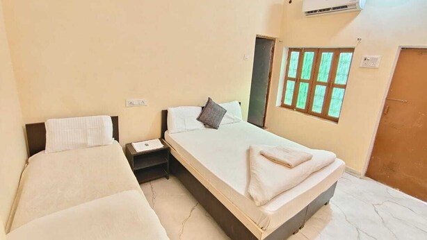 A hotel room with two single beds, a nightstand, an air conditioning unit, and a window with green foliage. - Hotel Shree (Varanasi)