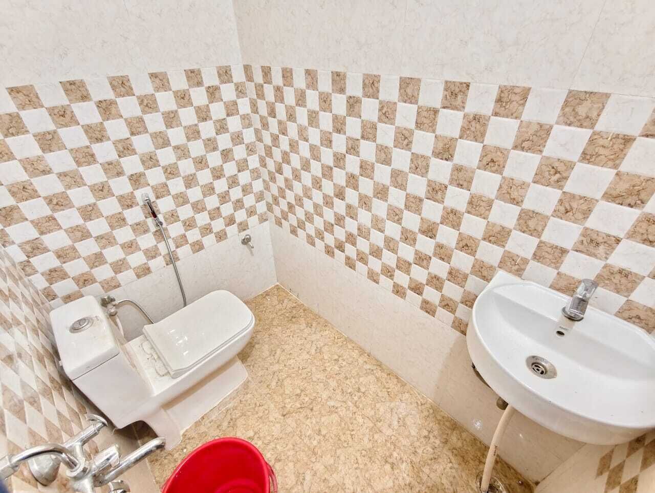 A bathroom with a checkered tile backsplash, a toilet, a sink, and a red bucket on the floor.