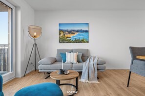 A modern living room with a sofa, a coffee table, a floor lamp, and a painting of a coastal landscape. - Biarritz - 2nd Floor, Balcony, Harbor View, Washing Machine, Private Parking (Olpenitz)
